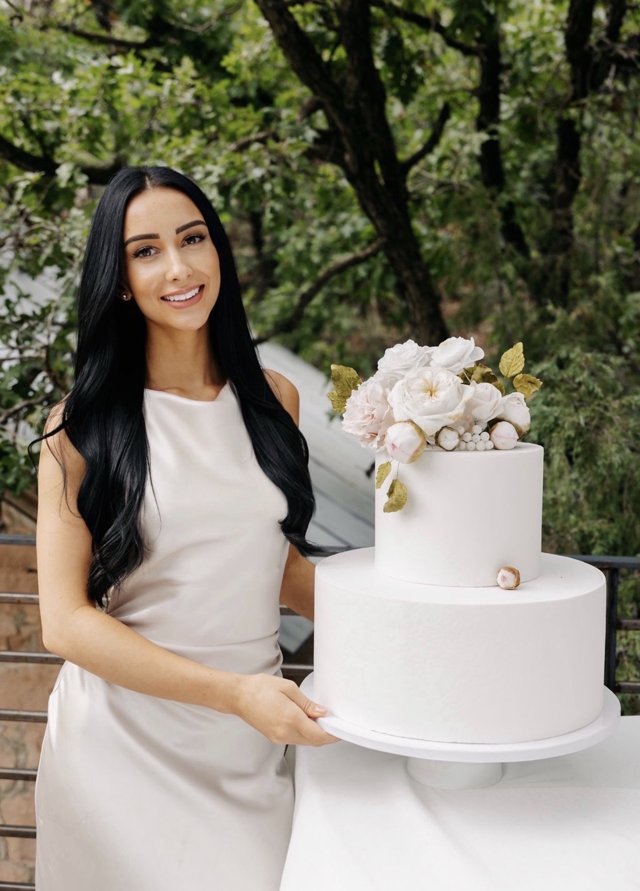 Magdalena smiling beside a two-tier white wedding cake topped with handmade sugar flowers