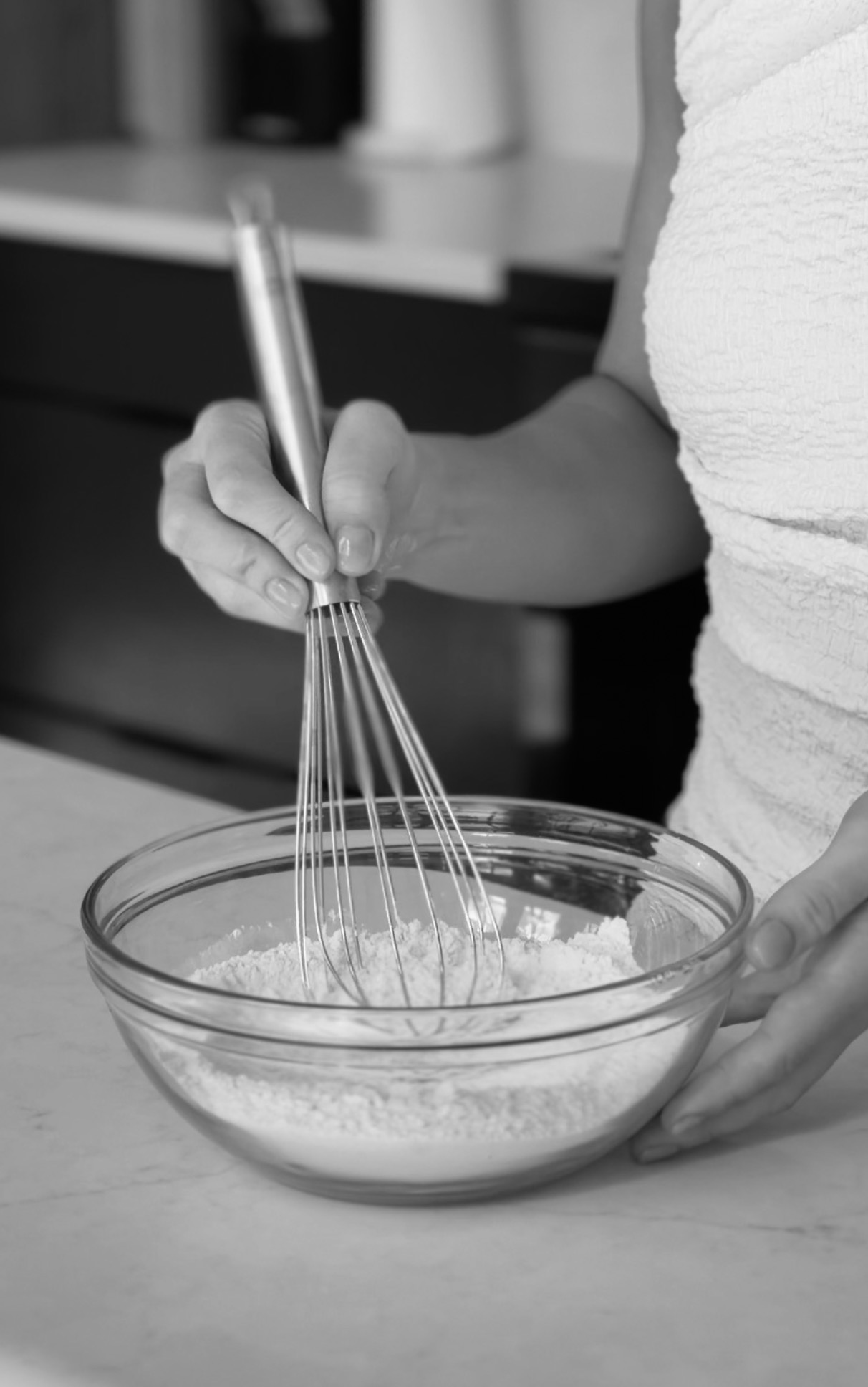 Black and white photograph of whisking flour in a glass bowl on the kitchen counter
