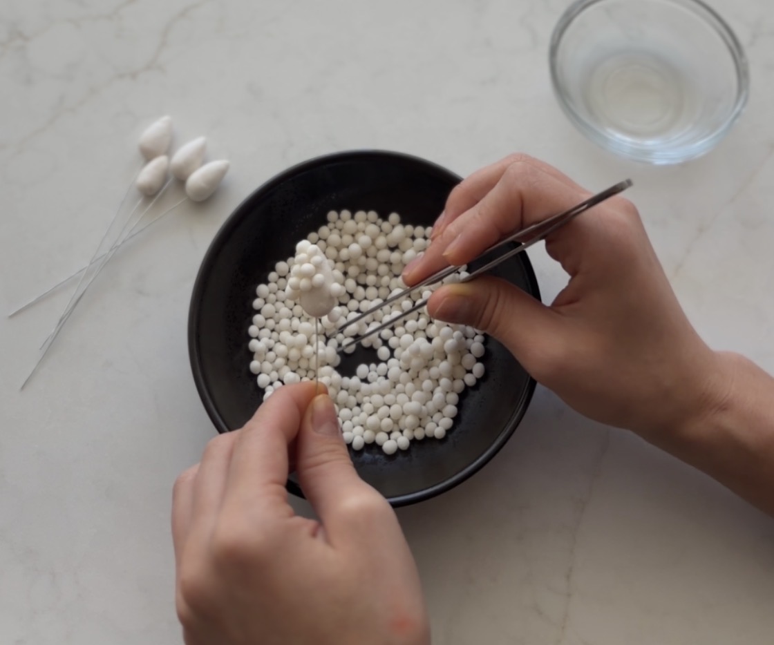 Overhead photograph of hands shaping a sugar flower with tweezers beside a bowl of sugar pearls