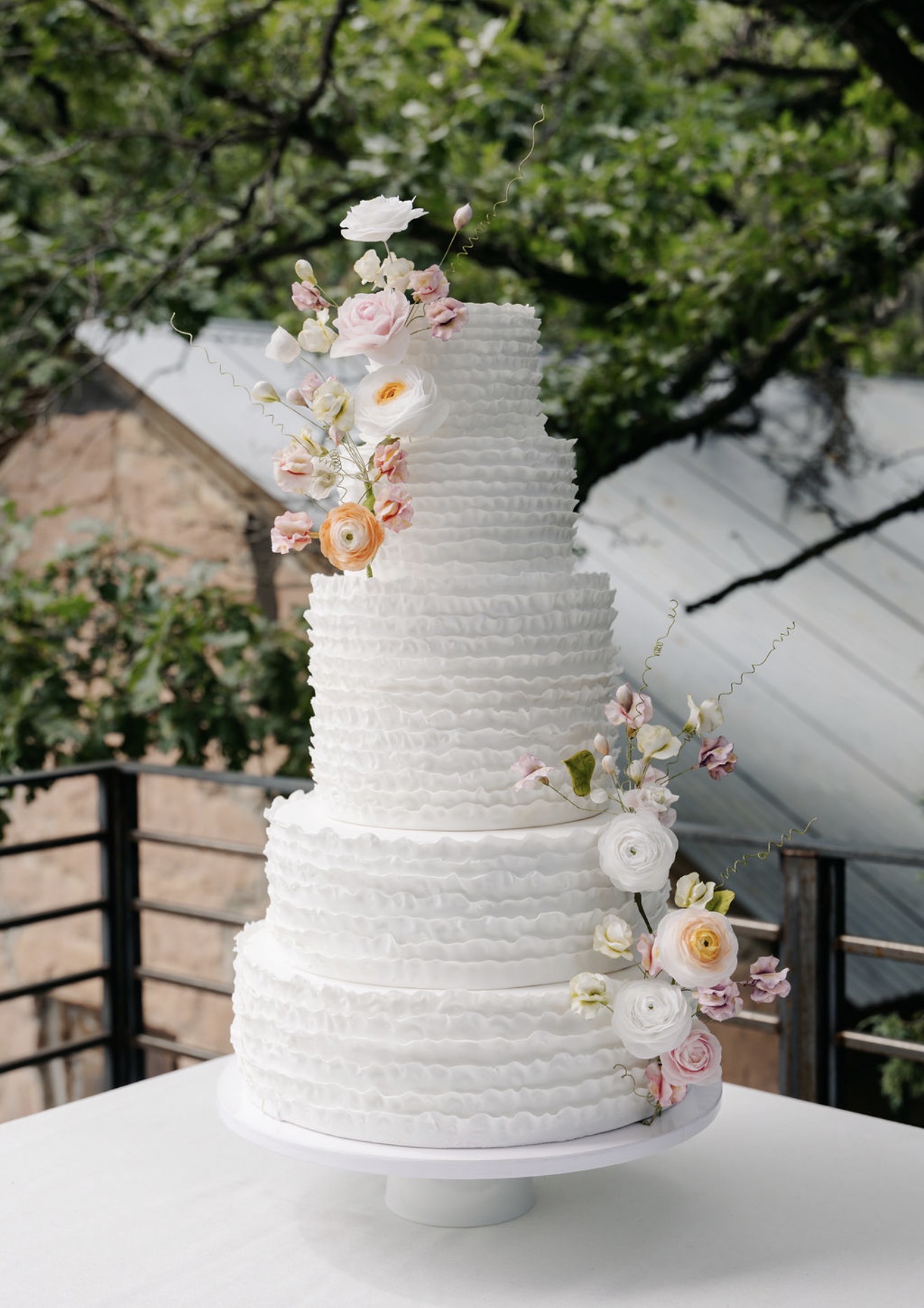 Tall ruffled-tier wedding cake with pastel ranunculus and sweet pea flowers, photographed outdoors